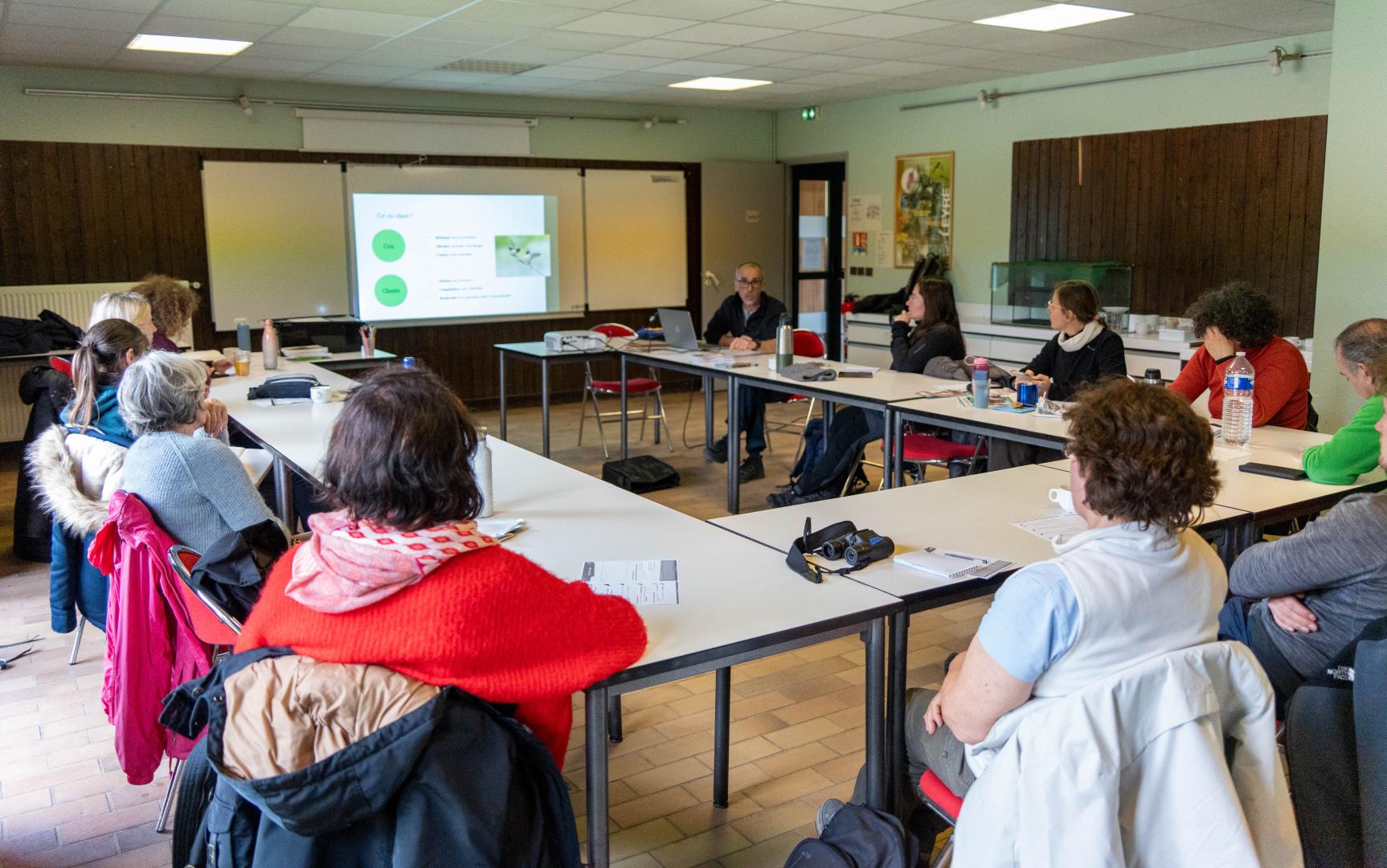 Des adultes assis en U écoutent un animateur qui présente un PowerPoint