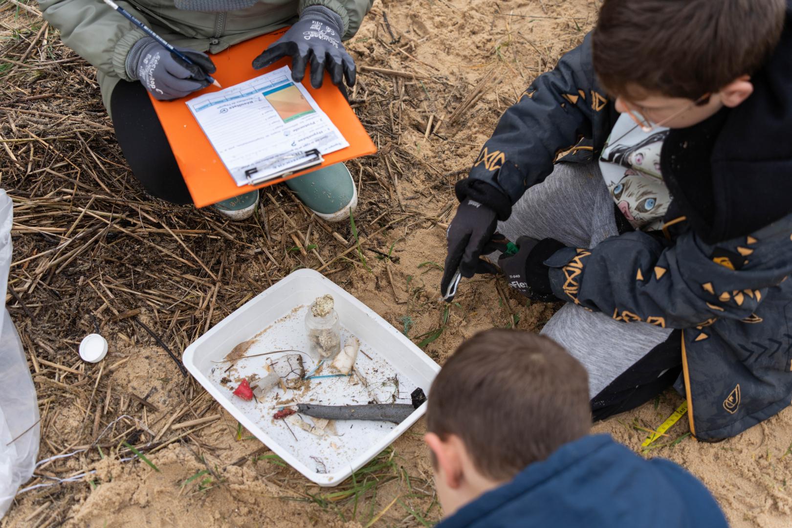 Sur le sable, des enfants sont penchés sur un bac qui contient des déchets plastiques, ils prennent des notes.